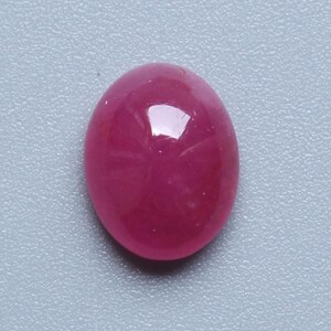 Könnte beinhalten: Ein polierter, oval geformter Rubin-Edelstein. Der Edelstein hat eine tiefe, gesättigte, rötlich-rosa Farbe mit einer glatten, reflektierenden Oberfläche. Der Edelstein liegt vor einem hellgrauen Hintergrund.