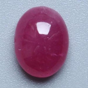 May include: A polished, oval-shaped ruby gemstone. The gem is a deep, saturated red color with a smooth, reflective surface. The stone is set against a light gray background.