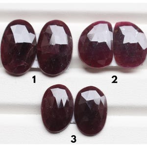 May include: Six faceted, deep red, oval-shaped gemstones arranged in three pairs, labeled 1, 2, and 3. Each stone features a polished, multi-faceted surface that reflects light, showcasing a dark crimson hue and a symmetrical cut for each pair.