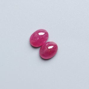 May include: Two oval-shaped, polished gemstones in a vibrant pink hue. The gems have a smooth, glossy surface, reflecting light. The stones are set against a light grey background.