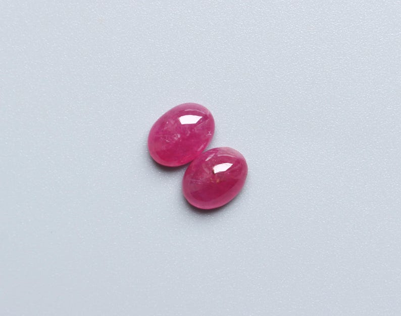 May include: Two oval-shaped, polished gemstones in a vibrant pink hue. The gems have a smooth, glossy surface, reflecting light. The stones are set against a light grey background.