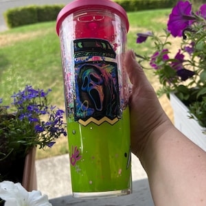 May include: A clear plastic tumbler with a bright pink lid and straw, featuring a vibrant green liquid base. The exterior displays a graphic of a VHS tape with a colourful, distorted ghost mask illustration and the text Scary Movies VHS.