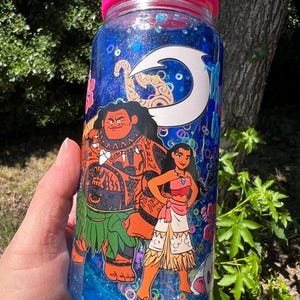 Puede incluir: Una botella de agua de pl&aacute;stico transparente con tapa rosa brillante, llena de l&iacute;quido azul con purpurina. El exterior presenta un estampado gr&aacute;fico de dos personajes animados, un hombre grande con tatuajes y una mujer, bajo un icono de anzuelo blanco.