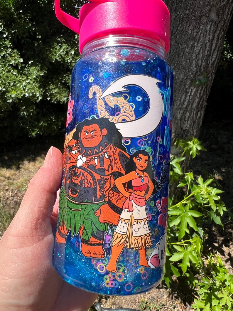 Puede incluir: Una botella de agua de pl&aacute;stico transparente con tapa rosa brillante, llena de l&iacute;quido azul con purpurina. El exterior presenta un estampado gr&aacute;fico de dos personajes animados, un hombre grande con tatuajes y una mujer, bajo un icono de anzuelo blanco.