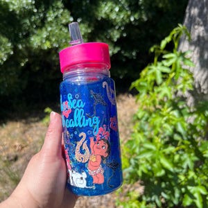 Puede incluir: Una botella de agua de pl&aacute;stico transparente con una tapa rosa brillante y una pajita transparente. La botella est&aacute; llena de l&iacute;quido azul y purpurina, con un personaje de dibujos animados, flores de hibisco, mantarrayas y el texto "The sea is calling" en azul.