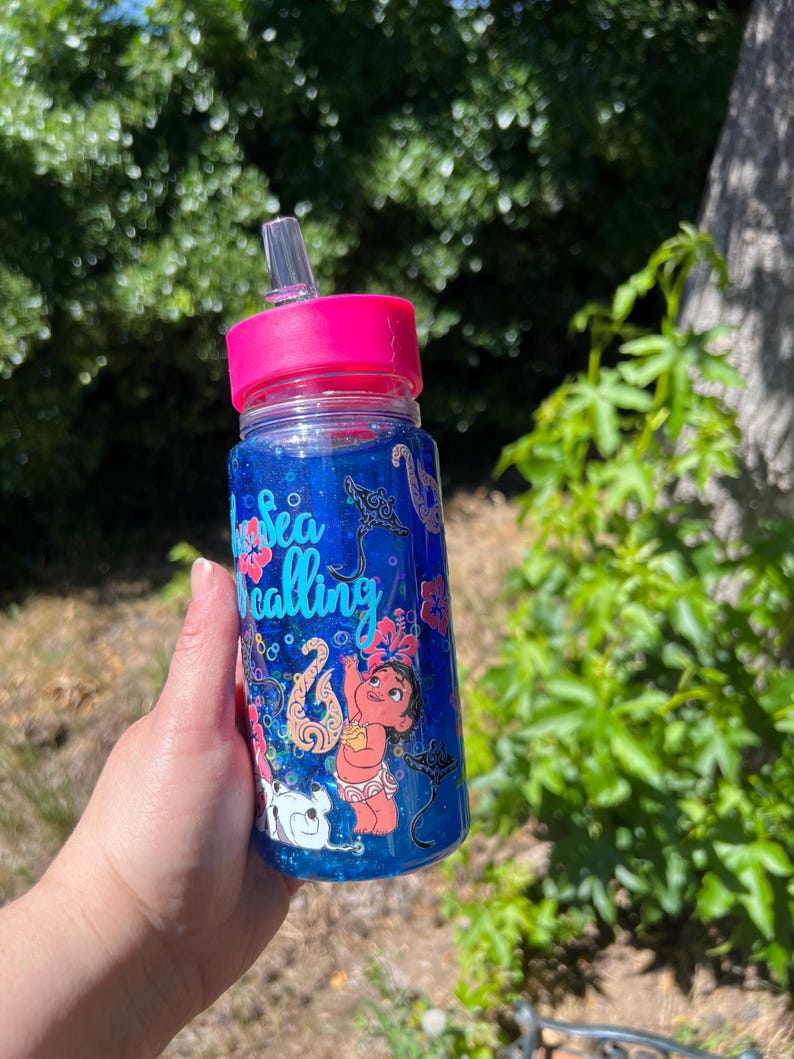 Puede incluir: Una botella de agua de pl&aacute;stico transparente con una tapa rosa brillante y una pajita transparente. La botella est&aacute; llena de l&iacute;quido azul y purpurina, con un personaje de dibujos animados, flores de hibisco, mantarrayas y el texto "The sea is calling" en azul.