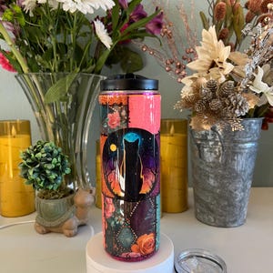 Puede incluir: Un vaso decorativo con tapa negra y tapa transparente extraíble. El vaso presenta un diseño con una silueta de gato negro sobre un cielo estrellado, rodeado de motivos florales y geométricos. El vaso mide aproximadamente 20 cm de alto.