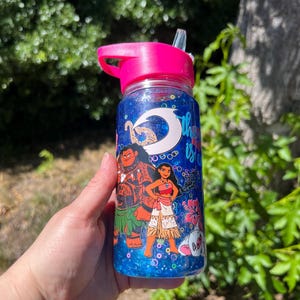 Puede incluir: Una botella de agua de pl&aacute;stico transparente con tapa y pajita de color rosa brillante, llena de l&iacute;quido azul con purpurina. El exterior presenta gr&aacute;ficos coloridos de dos personajes animados, un gancho blanco y flores de hibisco rosas.