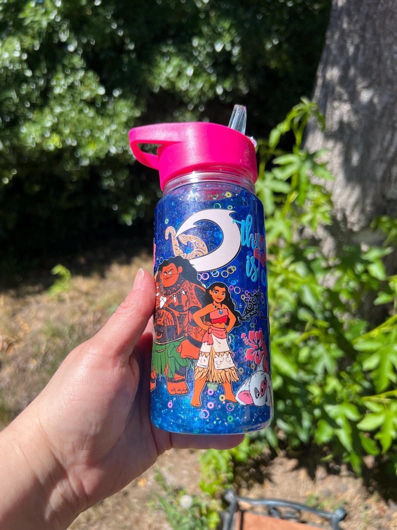 Puede incluir: Una botella de agua de pl&aacute;stico transparente con tapa y pajita de color rosa brillante, llena de l&iacute;quido azul con purpurina. El exterior presenta gr&aacute;ficos coloridos de dos personajes animados, un gancho blanco y flores de hibisco rosas.