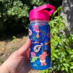 Puede incluir: Una botella de agua de pl&aacute;stico transparente con tapa y pajita de color rosa brillante, llena de l&iacute;quido azul con purpurina. El exterior presenta calcoman&iacute;as coloridas de un gallo, una raya, flores de hibisco, patrones tribales y un ni&ntilde;o.