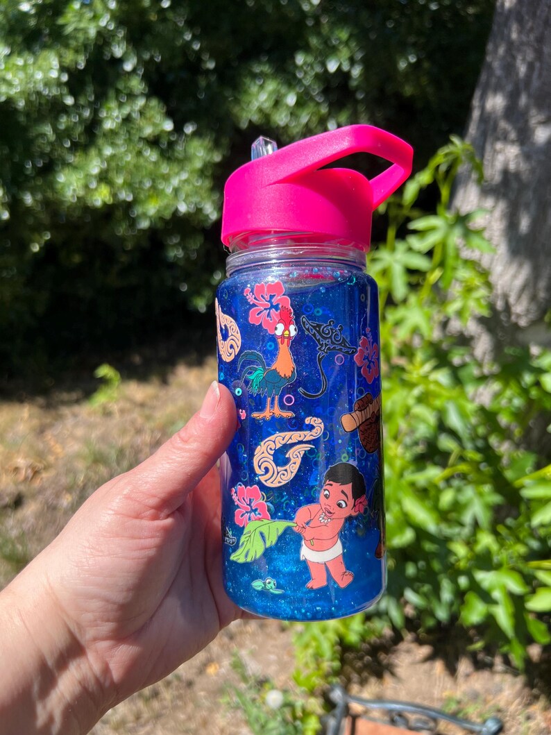 Puede incluir: Una botella de agua de pl&aacute;stico transparente con tapa y pajita de color rosa brillante, llena de l&iacute;quido azul con purpurina. El exterior presenta calcoman&iacute;as coloridas de un gallo, una raya, flores de hibisco, patrones tribales y un ni&ntilde;o.