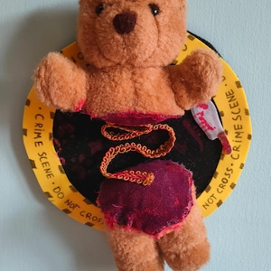 Pode incluir: Um urso de peluche castanho separado em duas peças, ligadas por uma fita metálica dourada. O urso está montado num sinal circular amarelo com o texto preto CRIME SCENE DO NOT CROSS. Um pequeno gráfico de uma faca branca está anexado.