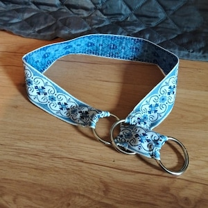Può includere: Una cintura in tessuto con un motivo floreale blu e bianco e un design geometrico ripetitivo. La cintura è fissata con due anelli metallici circolari color argento a un'estremità, attraverso i quali il cinturino è passato e fissato.