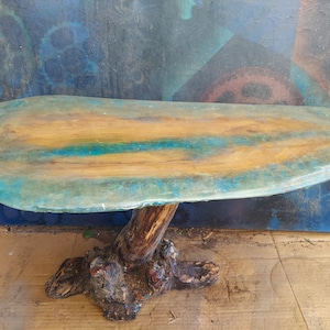 May include: A live edge table featuring a natural wood slab top with a central blue resin inlay. The table is supported by a single, thick, gnarled wooden tree root base that extends into a wide, textured foundation on the floor.
