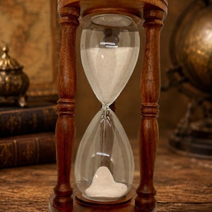 Könnte beinhalten: Eine Sanduhr mit einem dunkelbraunen Holzrahmen und gedrechselten Säulen, die eine kreisförmige Ober- und Unterseite verbinden. Der klare Glaskolben enthält feinen weißen Sand, der teilweise aus der oberen Kammer in die untere Kammer fließt.