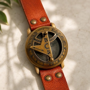 May include: A brass sundial compass with a brown leather strap featuring two metal rivets on each side. The circular brass casing displays Roman numerals and the text Ross London, with a central gnomon engraved with a sun and moon symbol.
