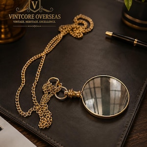 May include: A gold-toned magnifying glass with a circular lens and a matching gold-toned metal chain. The magnifying glass features a short handle attached to a circular frame. The text VINTCORE OVERSEAS VINTAGE. HERITAGE. EXCELLENCE. is visible.