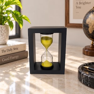 May include: An hourglass with a black rectangular frame and bright yellow sand. The glass bulbs are connected by a narrow neck, allowing the sand to flow from the top to the bottom bulb. The frame is solid black with a smooth, matte finish.