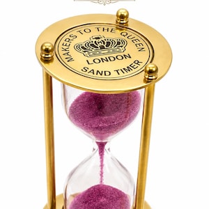 Może przedstawiać: Mosiężna klepsydra z okrągłą górną płytką wygrawerowaną napisem MAKERS TO THE QUEEN LONDON SAND TIMER oraz ikoną korony. Klepsydra zawiera żywy fioletowy piasek przepływający między dwiema przezroczystymi szklanymi bańkami podtrzymywanymi prętami.
