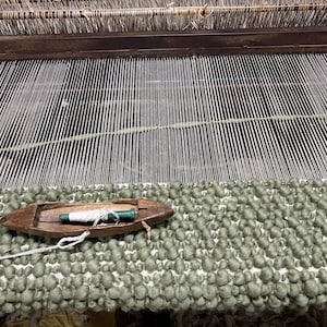 May include: A wooden weaving shuttle with a green and white bobbin rests on a textured sage green woven fabric. The shuttle is positioned on a loom with vertical white warp threads visible in the background, showing the process of textile creation.