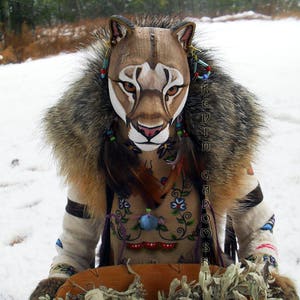 Puede incluir: Una persona con un abrigo marrón y blanco con ribete de piel, con una máscara de puma pintada, sosteniendo una cesta tejida llena de salvia seca. La persona está adornada con cuentas y plumas. El fondo es un paisaje nevado.