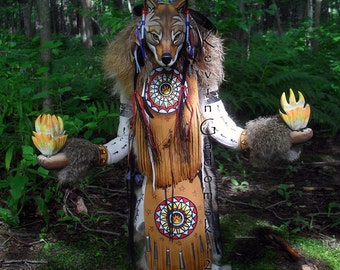 May include: A wooden Native American style doll with a wolf mask, feathers, and fringe. The doll is holding two orange flame-like objects in its hands. The doll is standing in a forest setting.