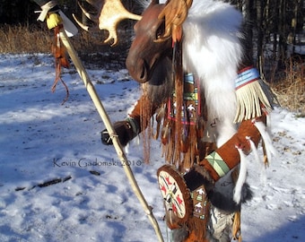 May include: A handcrafted figurine featuring a moose head with antlers, wearing brown leather clothing with fringe, white fur trim, and colorful geometric patterns. It holds a wooden staff topped with a bald eagle figure. Text reads Kevin Gadomski 2010.