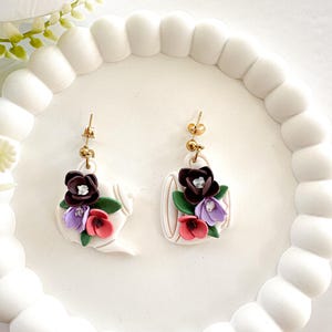May include: A pair of white teapot-shaped earrings with gold-colored posts. Each earring features a cluster of colorful flowers in shades of burgundy, purple, and pink, with green leaves. The earrings are displayed on a white, scalloped tray.