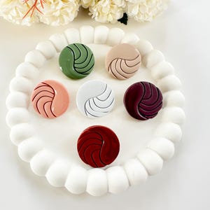 Op de afbeelding: Een verzameling van zes ronde decoratieve objecten in verschillende kleuren, waaronder groen, beige, roze, wit en bordeaux. Elk object heeft een gebogen, golfachtig ontwerp. Ze zijn gerangschikt op een witte, parelachtige tray, met witte bloemen op de achtergrond.