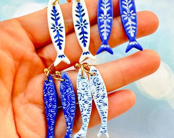May include: Handmade fish-shaped earrings in blue and white. The earrings feature intricate floral patterns and gold-colored ear wires. The fish designs are in various shades of blue and white, with a glossy finish. A unique and artistic accessory.