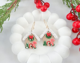 May include: A pair of festive gingerbread house earrings with gold-colored hooks. Each earring features a tan house with white frosting, a red bow, a green wreath, and a candy cane. The earrings are displayed on a white, cloud-shaped plate, with red berries and green sprigs in the background.