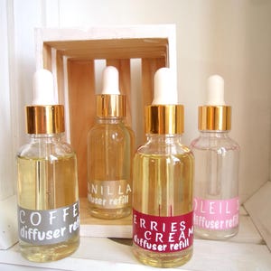 Può includere: Quattro flaconi di ricarica per diffusore in vetro trasparente con tappi dorati e contagocce bianchi. Le bottiglie sono etichettate con i profumi: Caffè, Vaniglia, Crema di Frutti di Bosco e Soleil. Esposti su una mensola di legno bianca.