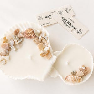 May include: Two seashell-shaped candles with decorative stones and seashells. The candles are white and have a textured edge. The image also includes two small tags with the text "Salt Kissed Ocean Breeze".