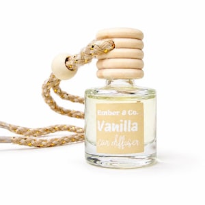 以下が含まれることがあります： 木製のキャップと「Vanilla car diffuser」と書かれたベージュ色のラベルが付いた、透明なガラス製の車用芳香剤ボトル。ボトルはベージュと金のロープで吊り下げられています。