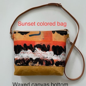 May include: A sunset-colored canvas bag with a brown leather strap and a waxed canvas bottom. The bag has a colorful abstract design with black, orange, and white paint.