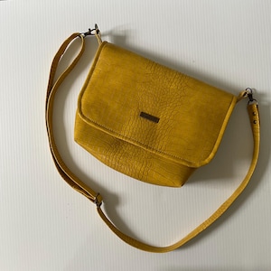 May include: Mustard yellow crossbody bag with a textured, crocodile-like pattern. The bag features a long, adjustable strap and a rectangular flap closure. A small, rectangular brown accent is centered on the flap.