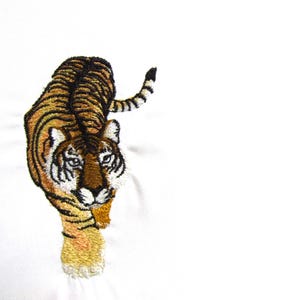 May include: Embroidered tiger design in shades of brown, black, and gold. The tiger is depicted in a walking pose, with its head turned towards the viewer. The embroidery is detailed, with visible stitching and texture. The background is white.