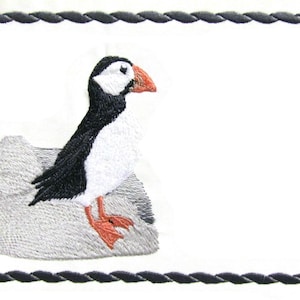 May include: Embroidered image of a puffin bird perched on a gray rock. The bird has black and white feathers, an orange beak and feet. The image is framed with a black rope-like border on a white background.