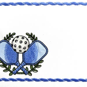 May include: Embroidered white fabric with a blue rope-like border. The center features a pickleball design with two crossed blue paddles, a white ball, and green laurel leaves. A sports-themed item.