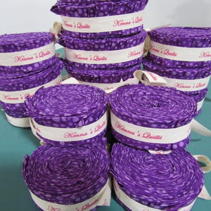 Peut inclure: Plusieurs rouleaux de tissu violet à pois blancs, entourés de bandes blanches avec l'inscription "Kenna's Quilts" en rose. Le tissu est roulé et attaché avec des rubans blancs, pour des projets de couture.