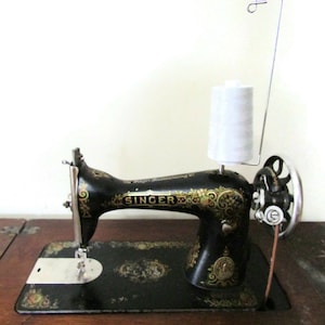 May include: A vintage Singer sewing machine, black with gold floral detailing, is displayed on a wooden surface. A spool of white thread is held by a wire stand. The Singer logo is clearly visible on the machine.
