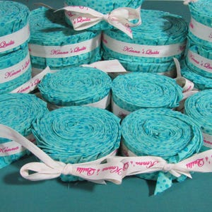 May include: Stacks of rolled turquoise fabric with white ribbon accents. The fabric has a polka dot pattern. The ribbon is printed with "Konna's Quilts" in pink script. Ideal for quilting projects.