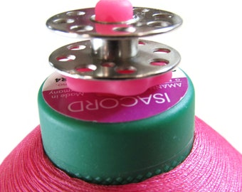 Può includere: Primo piano di una bobina di filo ISA CORD rosa con base verde e parte superiore argentata e rosa. L'etichetta recita "ISA CORD" e "Made in Germany". Il filo è ben avvolto e la bobina è su sfondo bianco.