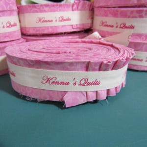 May include: Pink fabric rolls with white labels that read "Kenna's Quilts".
