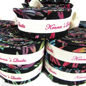 May include: Rolled fabric bundles with a colourful floral pattern on a black background. Each bundle is wrapped with a white band that reads "Kenna's Quilts" in pink script. The bundles are stacked, ideal for quilting projects.