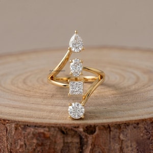 Può includere: Un anello in oro con quattro pietre trasparenti sfaccettate di forme diverse: un taglio a goccia in alto, un taglio ovale, un taglio princess quadrato e un taglio brillante rotondo in basso. La fascia ha un design a bypass con il testo AshauraJewels.