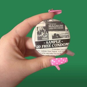 May include: A round button with a black and white design. The button features the text "- SAMPLE - 20 FREE CONDOMS" and advertisements for Trojan Magnum and Rough Rider condoms. The background is a solid green.