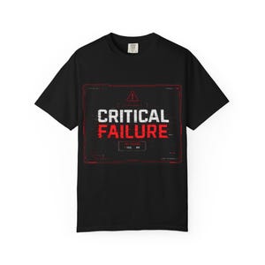 Op de afbeelding: Een zwart t-shirt met korte mouwen en een grafische print van een rood en wit digitaal interfacekader. De tekst CRITICAL FAILURE staat in het midden gedrukt, met een waarschuwingsdriehoek erboven en een vak met YES en NO opties eronder.