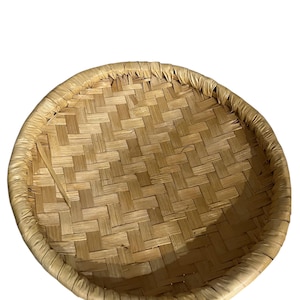May include: A round, woven basket with a natural, light brown color. The basket features a tightly woven pattern and a slightly raised rim. The texture of the basket is visible, showcasing the individual strands of material used in its construction.
