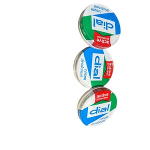 May include: Three round metal containers of Dial cream deodorant are stacked on a white background. Each container has a blue top with the brand name "dial" in white, a red section with "active protection", and a green rim.
